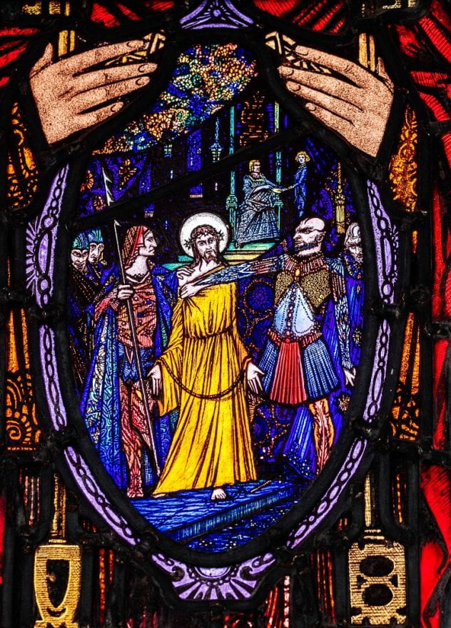 Stained glass window of the first of the fourteen stations of the cross showing Jesus being convicted. The image is on a shield held by St. Peter